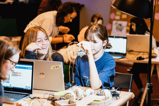 Zwei t&uuml;ftelnde M&auml;dchen sitzen lachend fasziniert vor einem Laptop und versuchen sich am Programmieren eines calliope mini Controllers.