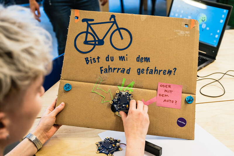 E! Eine Mentorin h&auml;lt ein Workshop-Ergebnis in den H&auml;nden: Ein St&uuml;ck Karton auf dem steht "Bist du mit dem Fahrrad gefahren oder mit dem Auto?" Ein darauf befestigter Mirkocontroller erm&ouml;glicht verschiedene Antwortm&ouml;glichkeiten.