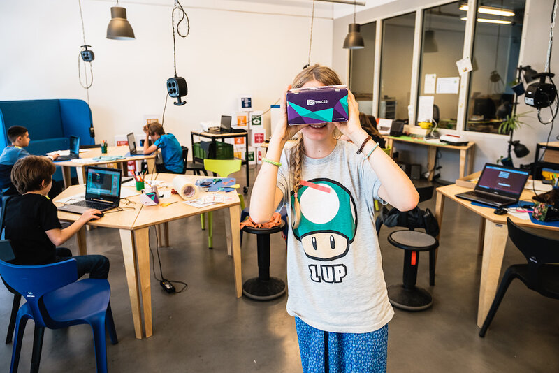 Blick ins GoodLab Berlin. Im Vordergrund hält ein Kind sich eine VR-Brille vor die Augen. Im Hintergrund sieht man Kinder, die mit Laptops und Technikzubehör arbeiten.