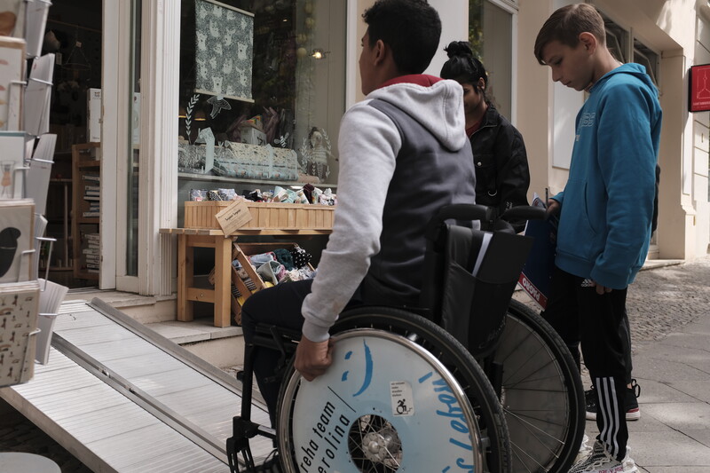 Ein Junge im Rollstuhl steht an der Rampe vor der Eingangst&uuml;r eines Ladengesch&auml;fts