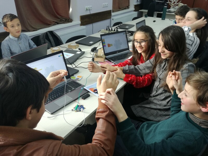 Kinder sitzen vor einem Laptop und halten sich an den H&auml;nden, um den Stromkreis des Mikrocontrollers zu schlie&szlig;en