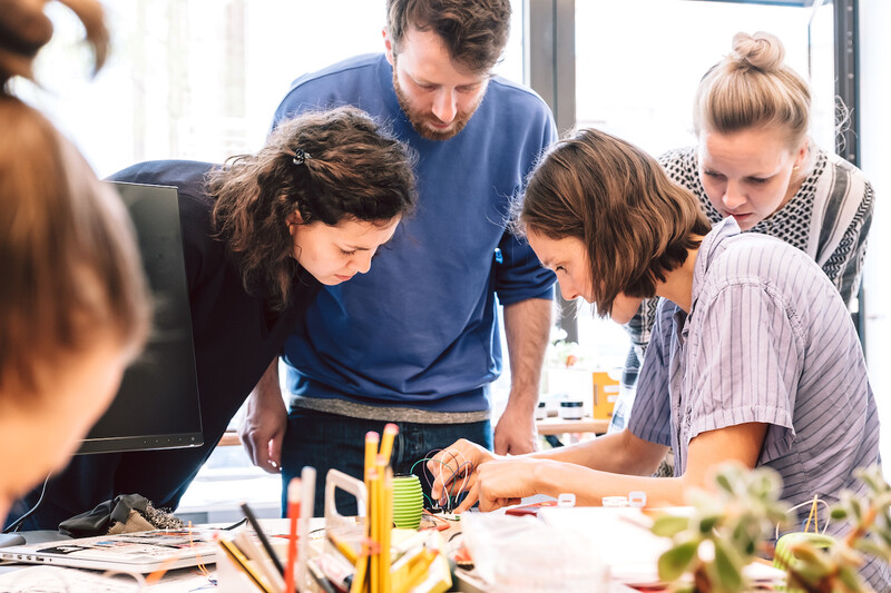 Eine Person steckt Kabel an eine Bluetooth Box und an einen Mikrocontroller. Drei Erwachsene stehen um den Tisch und schauen sich das genau an.