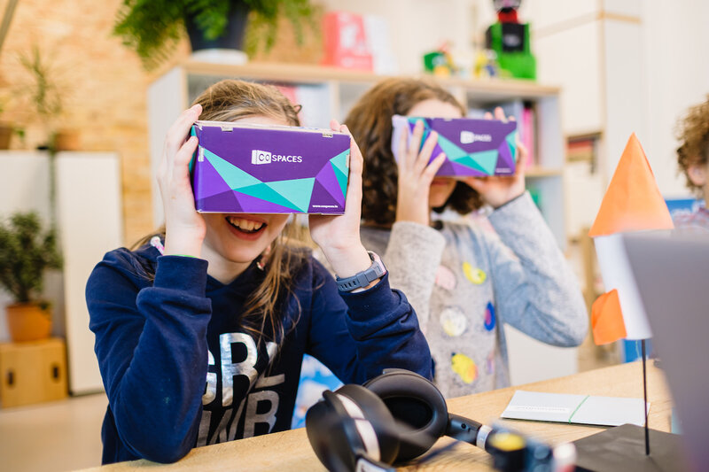 Zwei Kinder sitzen an einem Tisch und halten sich VR-Brillen vor ihr Gesicht.
