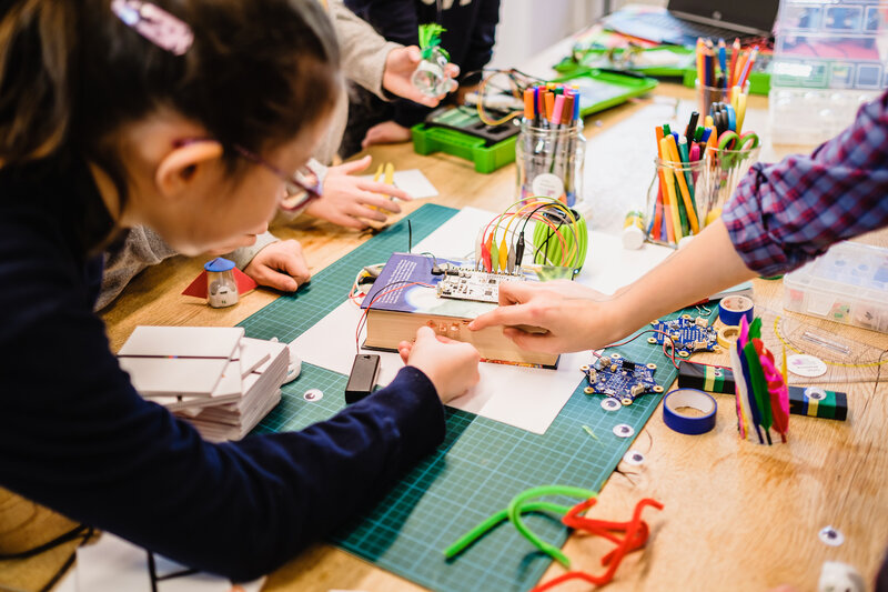 Ein Kind tüftelt mit Kabeln und einem Mikrocontroller. In der Mitte eines Tisches liegt ein Buch auf dem eine Leiterplatine mit vielen verschiedenen Kabeln liegt.