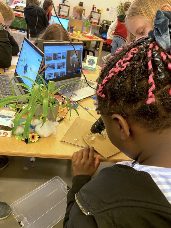 Ein Kind klebt mit einer Heißklebepistole ein Bauteil zusammen. Im Hintergund sieht man mehrere Kinder, Laptops und verschiedene Bauteile für Roboter.