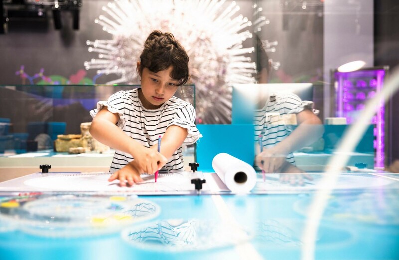 Ein junges M&auml;dchen probiert an einer Mitmachstation einen Spirographen aus, im Hintergrund sind Exponate des Futuriums zu sehen.